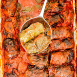 overhead shot of a serving spoon holding up two stuffed grape leaves above the pan of stuffed grape leaves