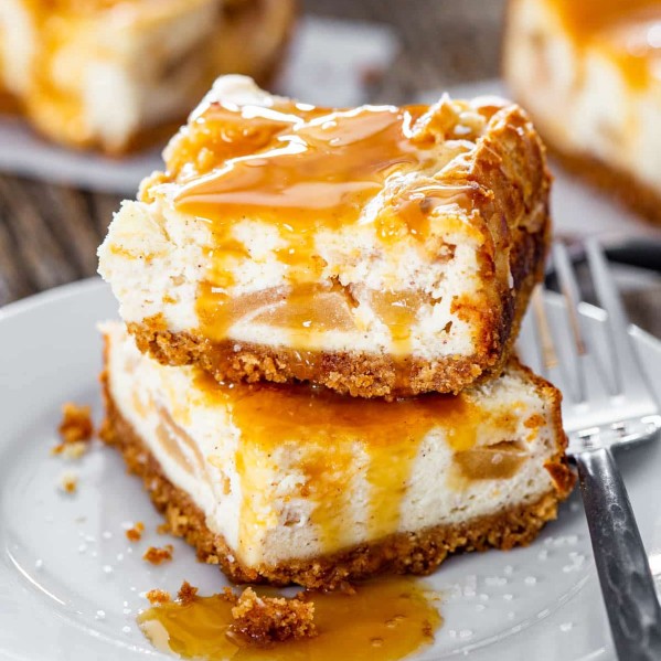 close up shot of two caramel apple pie cheesecake bars covered in caramel sauce stacked on top of one another on a plate