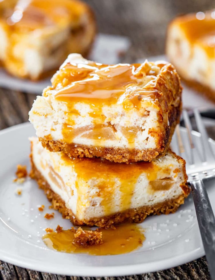 close up shot of two caramel apple pie cheesecake bars covered in caramel sauce stacked on top of one another on a plate