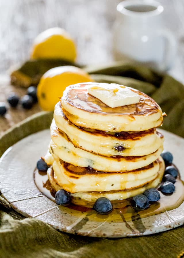a stack of ricotta pancakes drizzled with syrup, surrounded by blueberries, with a pad of butter on top