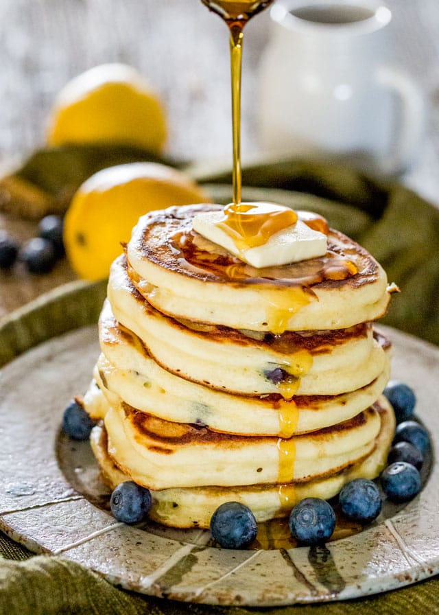 a stack of ricotta pancakes surrounded by blueberries with syrup being drizzled over them