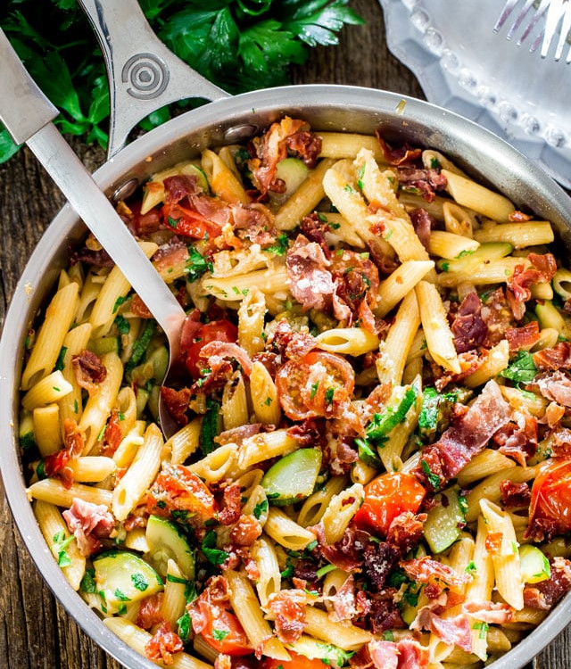 overhead shot of a serving spoon resting in a pot of penne with prosciutto tomatoes and zucchini