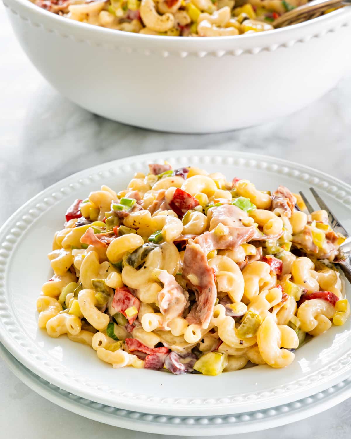 a white plate filled with macaroni salad with prosciutto with a big bowl in the background