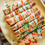 overhead shot of a stack of chicken enchilada taquitos drizzled with hot sauce and crema