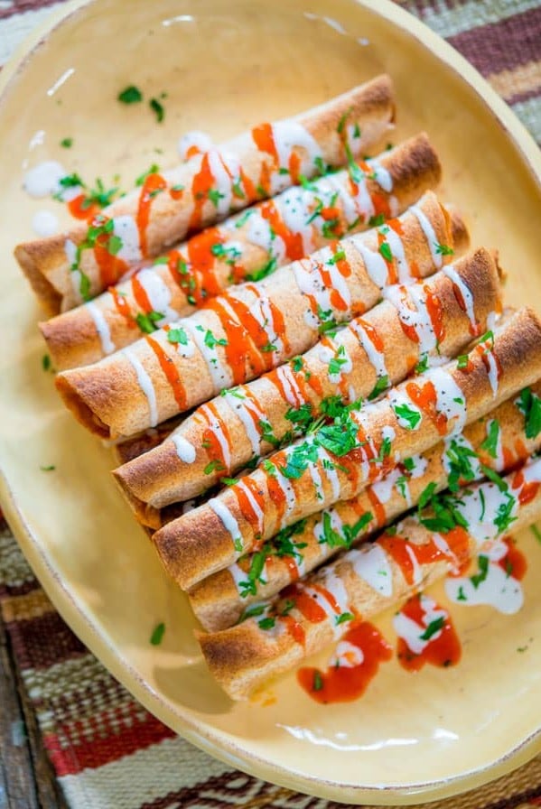 overhead shot of a stack of chicken enchilada taquitos drizzled with hot sauce and crema