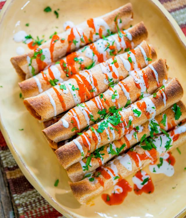overhead shot of a stack of chicken enchilada taquitos drizzled with hot sauce and crema