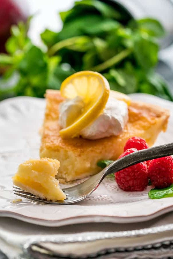 side view shot of a fork taking a bite of a slice of lemon chess pie topped with whipped cream and a slice of lemon on a plate garnished with fresh raspberries