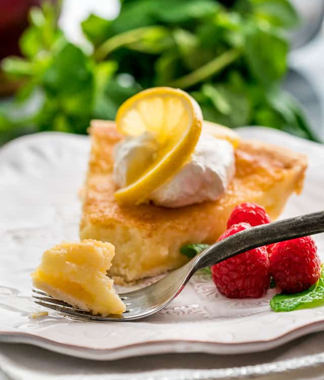 side view shot of a fork taking a bite of a slice of lemon chess pie topped with whipped cream and a slice of lemon on a plate garnished with fresh raspberries