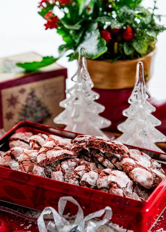 a cookie tin full of red velvet crinkle cookies with christmas decorations in the background