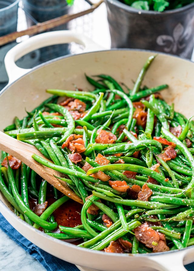 side view shot of green beans and bacon in a pan being stirred by a wooden spoon