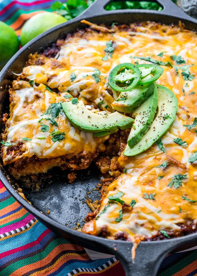 a skillet filled with cheesy casserole with a piece taken out topped with avocado and jalapeno