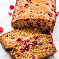 side view shot of cranberry quick bread with two slices cut. Fresh cranberries surround the loaf of bread