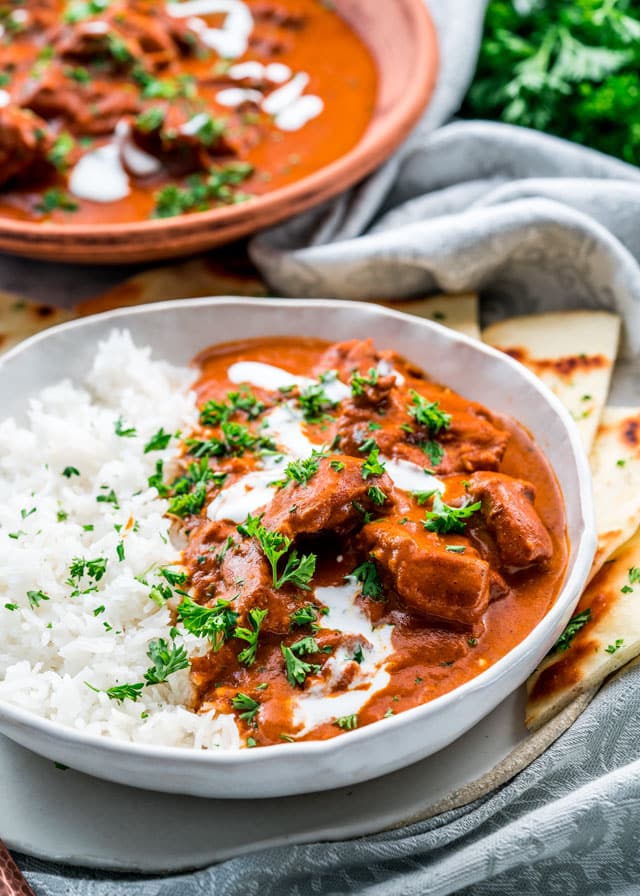 closeup of butter chicken and rice in a white bowl surrounded with naan