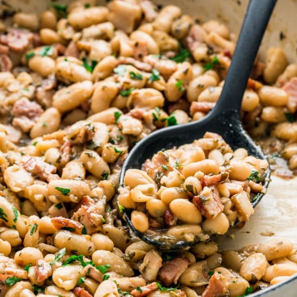 side view shot of a serving spoon taking a scoop of white beans and bacon from a pot