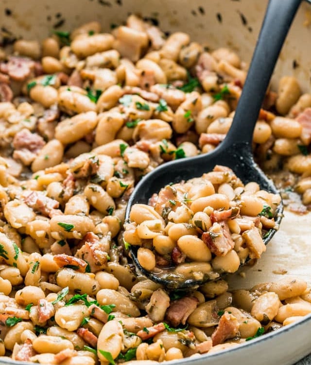 side view shot of a serving spoon taking a scoop of white beans and bacon from a pot