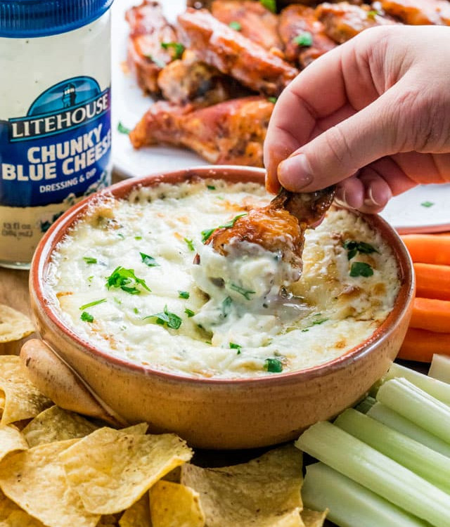 side view shot of a hand dipping a buffalo wing into the blue cheese dip. the bowl is surrounded by tortilla chips, carrot sticks and celery slices.
