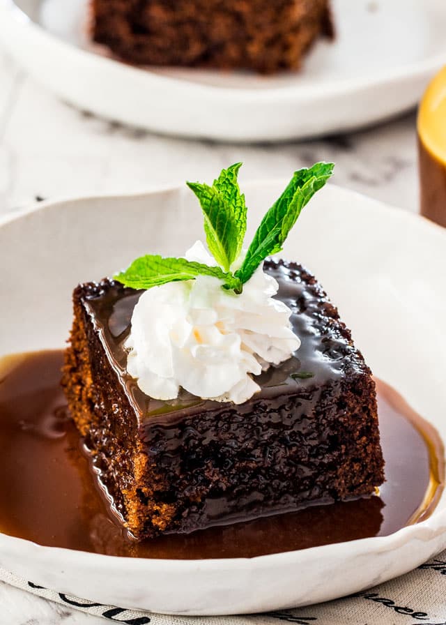 a piece of sticky toffee pudding in a white plate drenched with sticky toffee sauce and topped with whipped cream