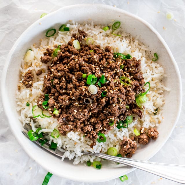  Korean Beef over rice in a white bowl garnished with sesame seeds and green onions in a white bowl with a fork