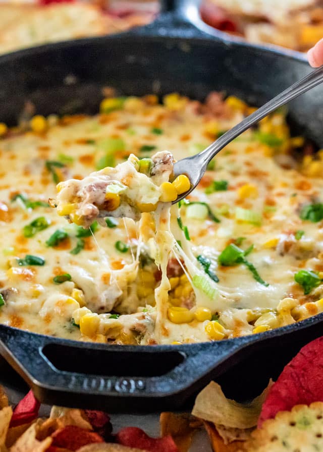 A fork loaded up with a bite of bacon corn dip with cheese stretching back into the skillet