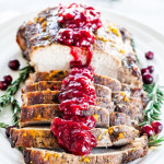 sliced pork roast topped with cranberry sauce on a plate garnished with rosemarry and cranberries