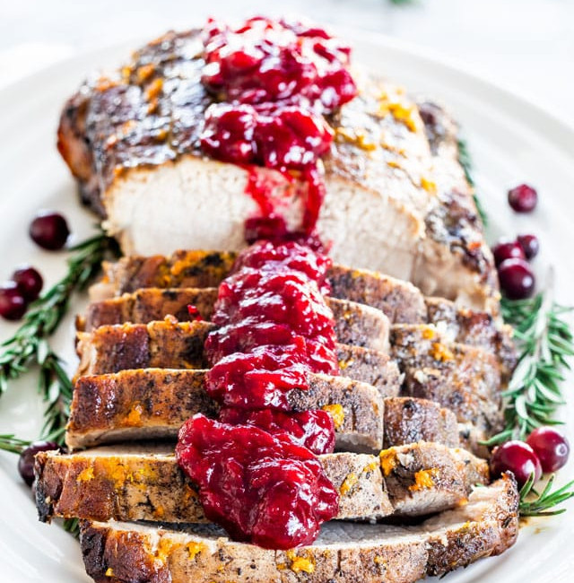 sliced pork roast topped with cranberry sauce on a plate garnished with rosemarry and cranberries