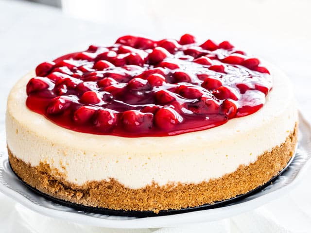 cheesecake on a white cake platter topped with cherry pie filling