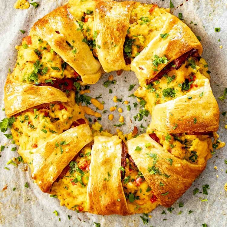 Overhead view of golden crescent breakfast ring filled with scrambled eggs, bacon, cheese, and peppers on parchment paper.