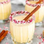 glasses of eggnog in glasses rimmed with chocolate and crushed candy cane pieces, garnished with cinnamon sticks.
