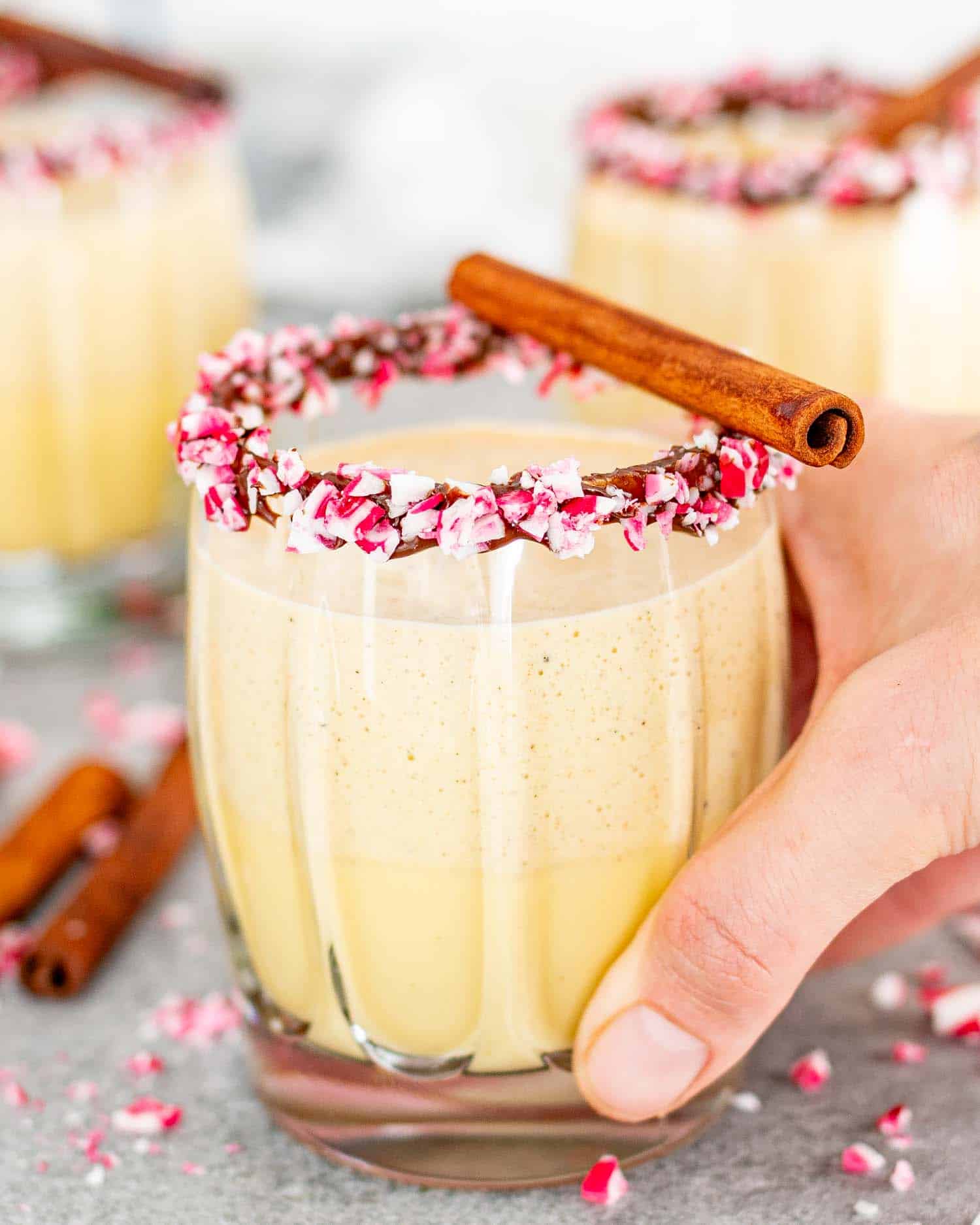 glasses of eggnog in glasses rimmed with chocolate and crushed candy cane pieces, garnished with cinnamon sticks.