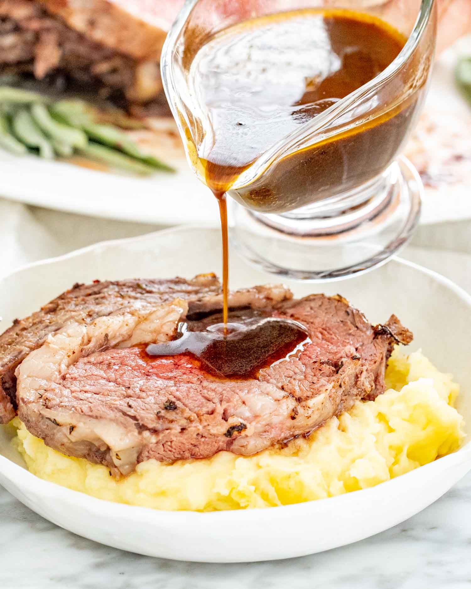 pouring gravy on a piece of prime rib roast that sits atop a bed of mashed potatoes.