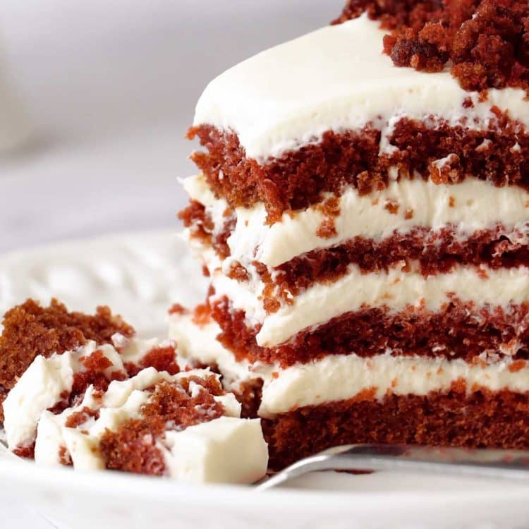 Slice of red velvet cake with cream cheese frosting showing moist layers and rich red crumb on a white plate.