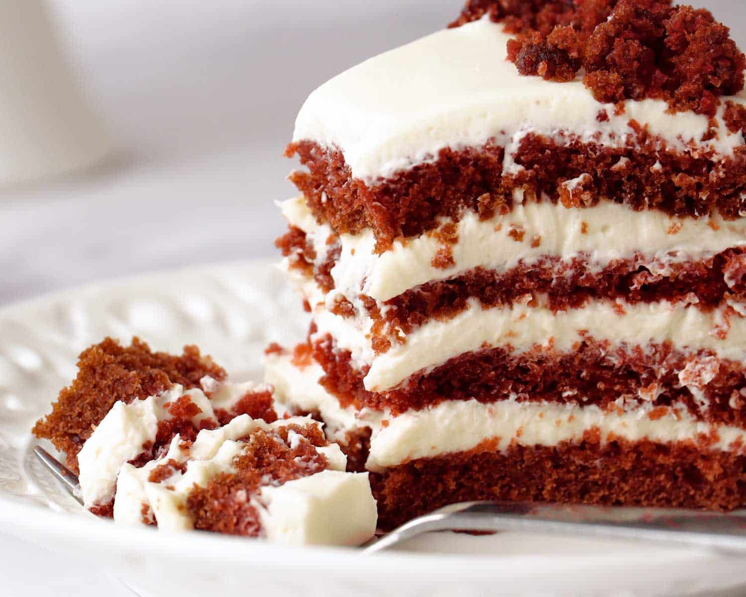 Slice of red velvet cake with cream cheese frosting showing moist layers and rich red crumb on a white plate.