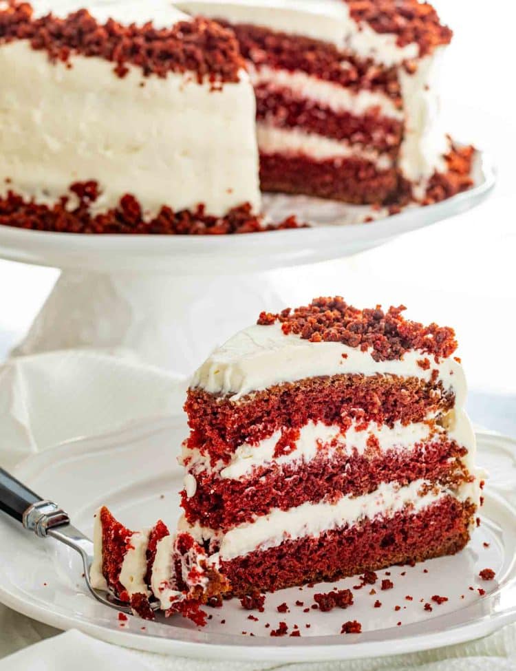 Red velvet cake slice served on white plate with fork showing soft texture and creamy frosting.