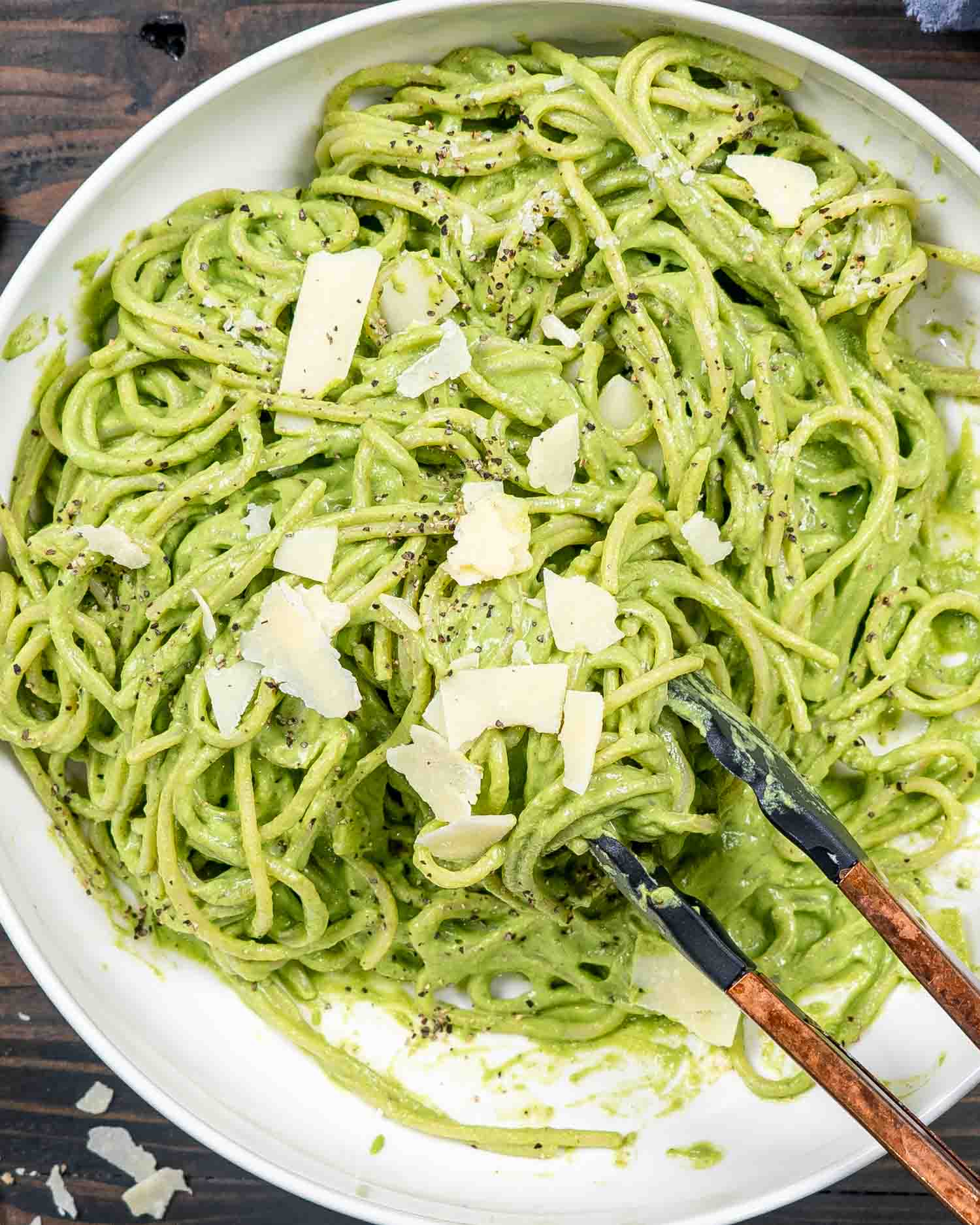 a white bowl with freshly made creamy avocado spinach pasta with a pair of tongs and garnished with parmesan.