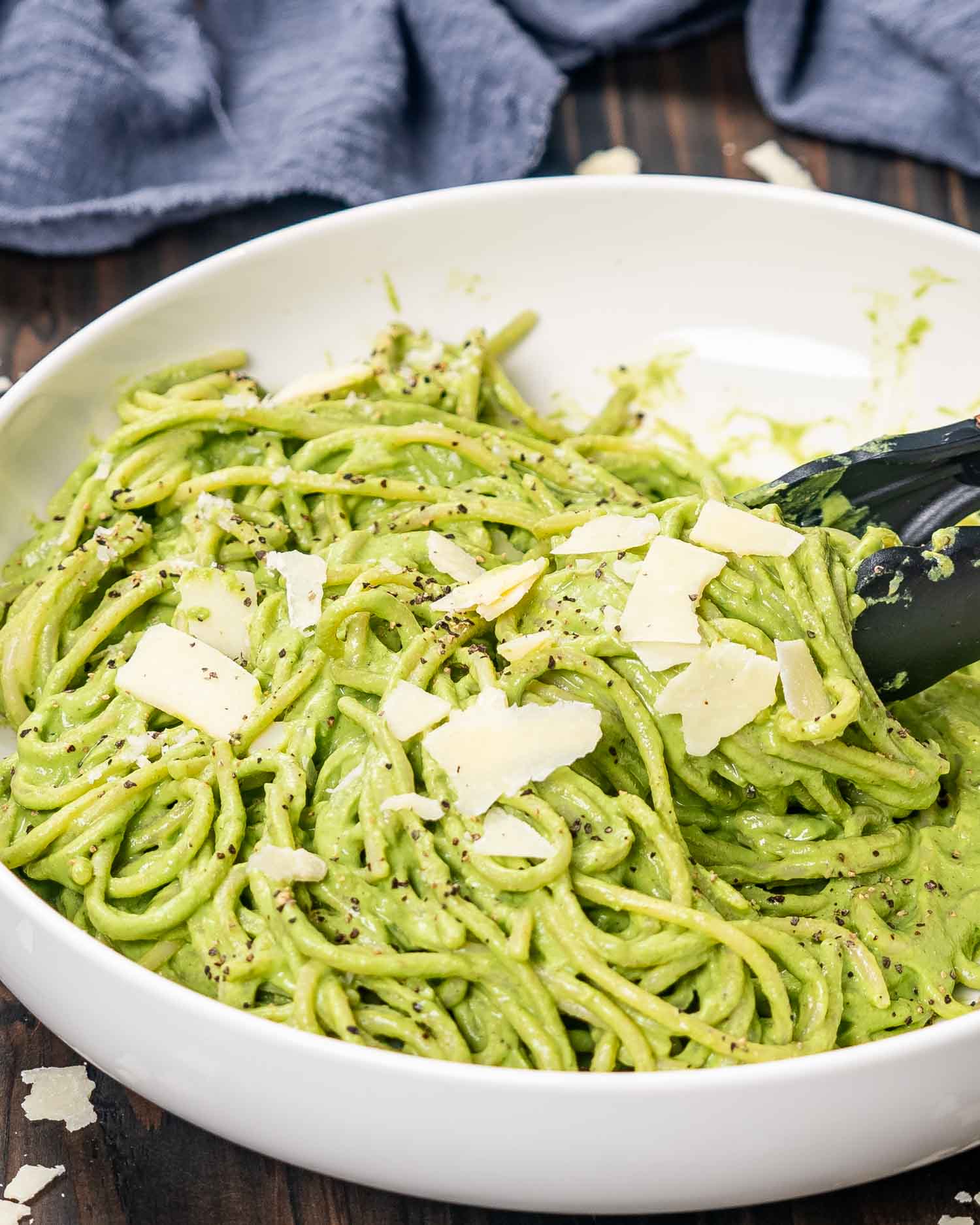 a white bowl with freshly made creamy avocado spinach pasta with a pair of tongs and garnished with parmesan.