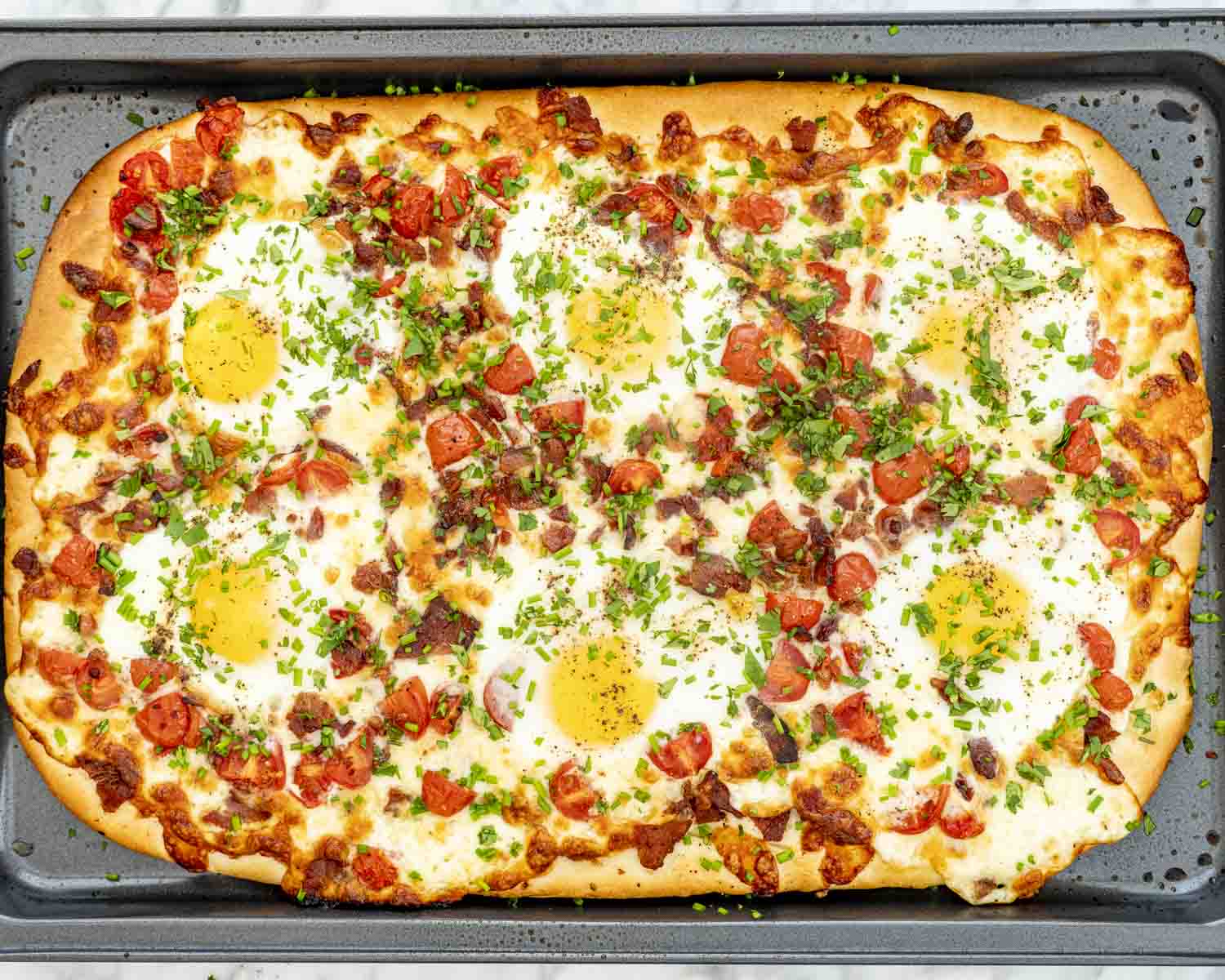Large sheet pan breakfast pizza baked with eggs, tomatoes, bacon, mozzarella, and fresh parsley, ready to slice and serve.