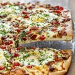 Slice of cheesy breakfast pizza being lifted, showing gooey melted cheese, bacon, egg, tomatoes, and fresh herbs.