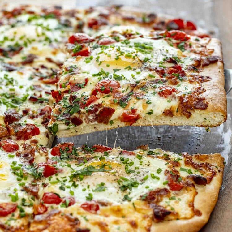 Slice of cheesy breakfast pizza being lifted, showing gooey melted cheese, bacon, egg, tomatoes, and fresh herbs.