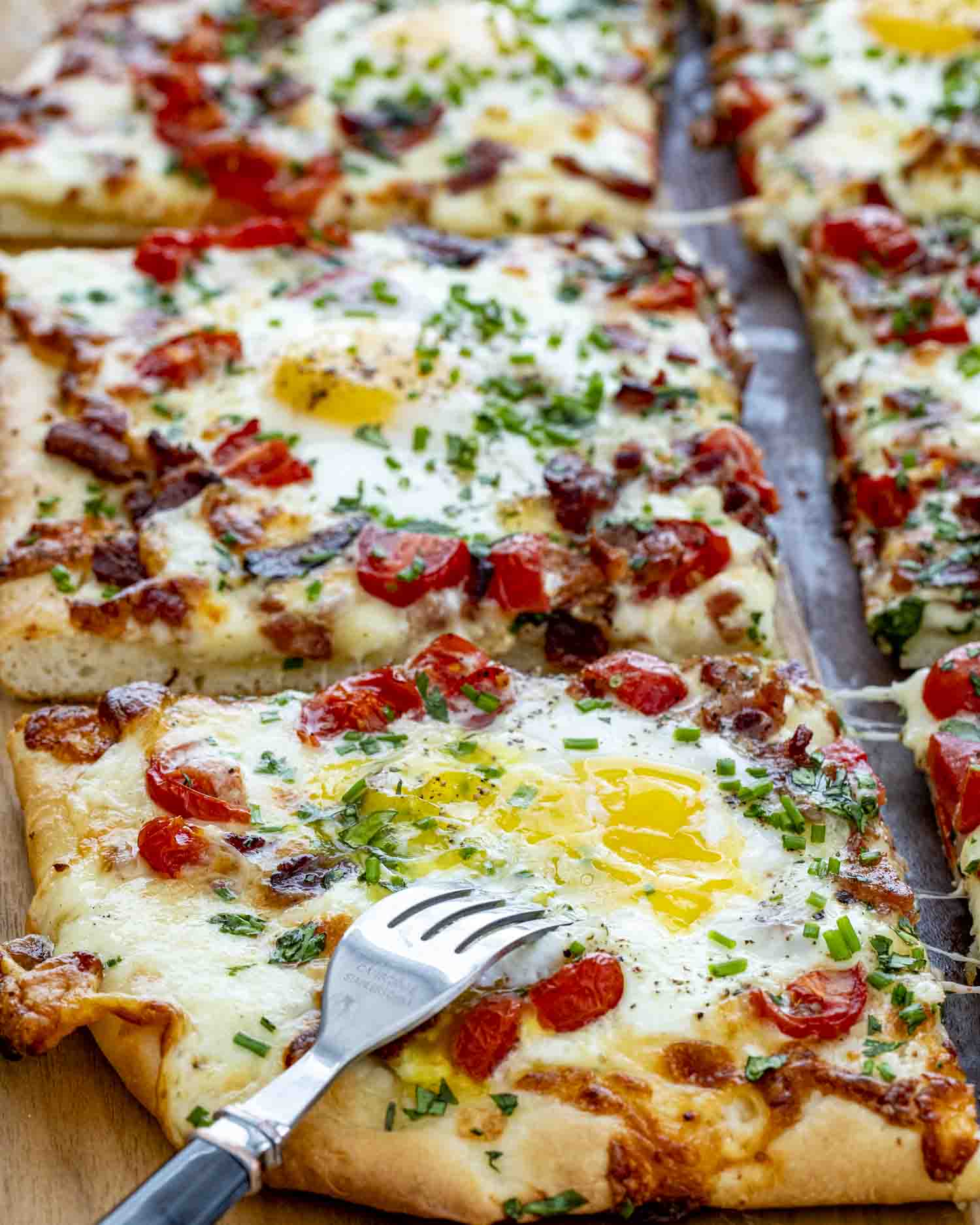 Close-up of cheesy breakfast pizza slice topped with egg, bacon, tomatoes, and herbs, cut with a fork.