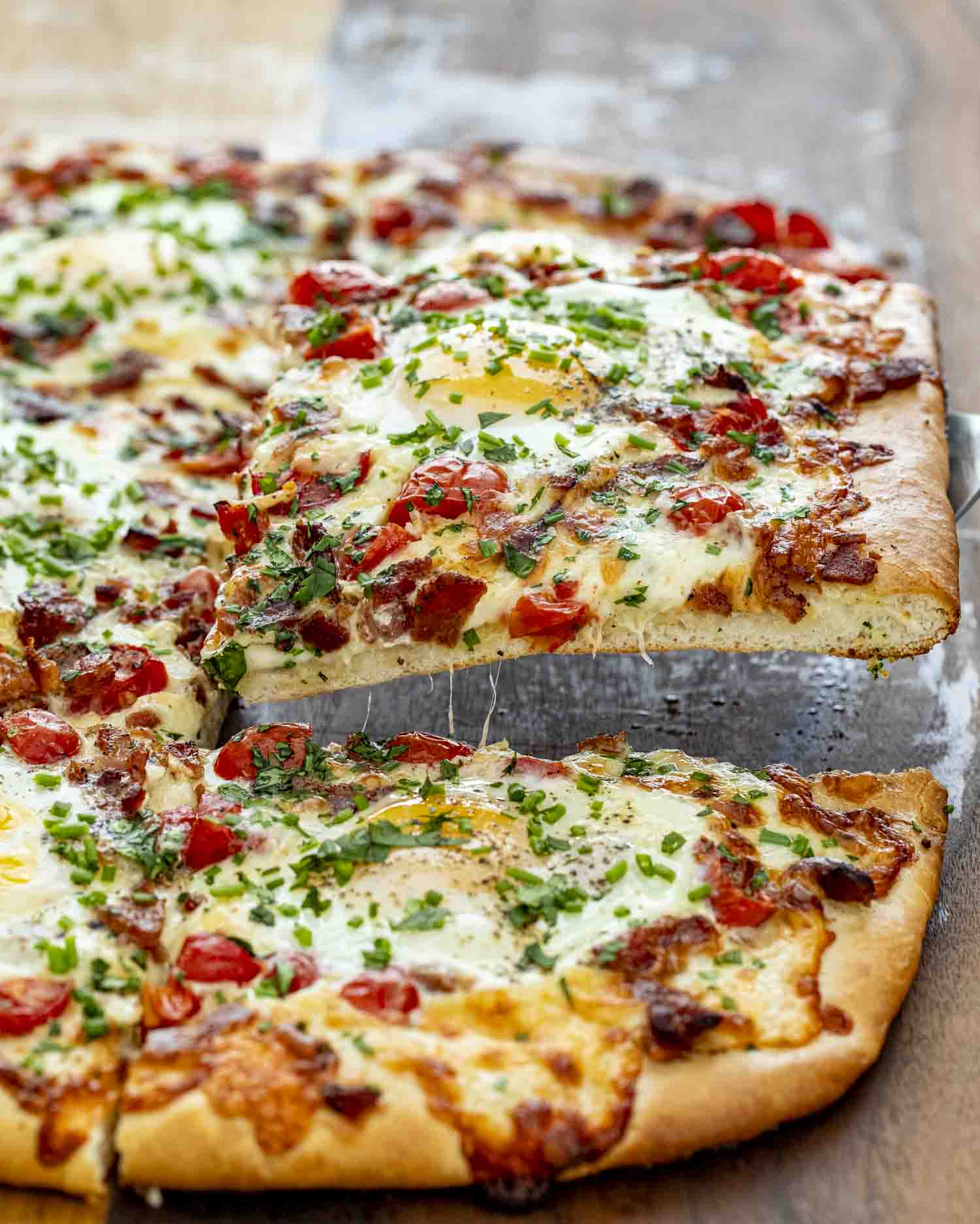Slice of cheesy breakfast pizza being lifted, showing gooey melted cheese, bacon, egg, tomatoes, and fresh herbs.
