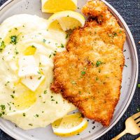 a chicken schnitzel on a plate with mashed potatoes and lemon wedges.