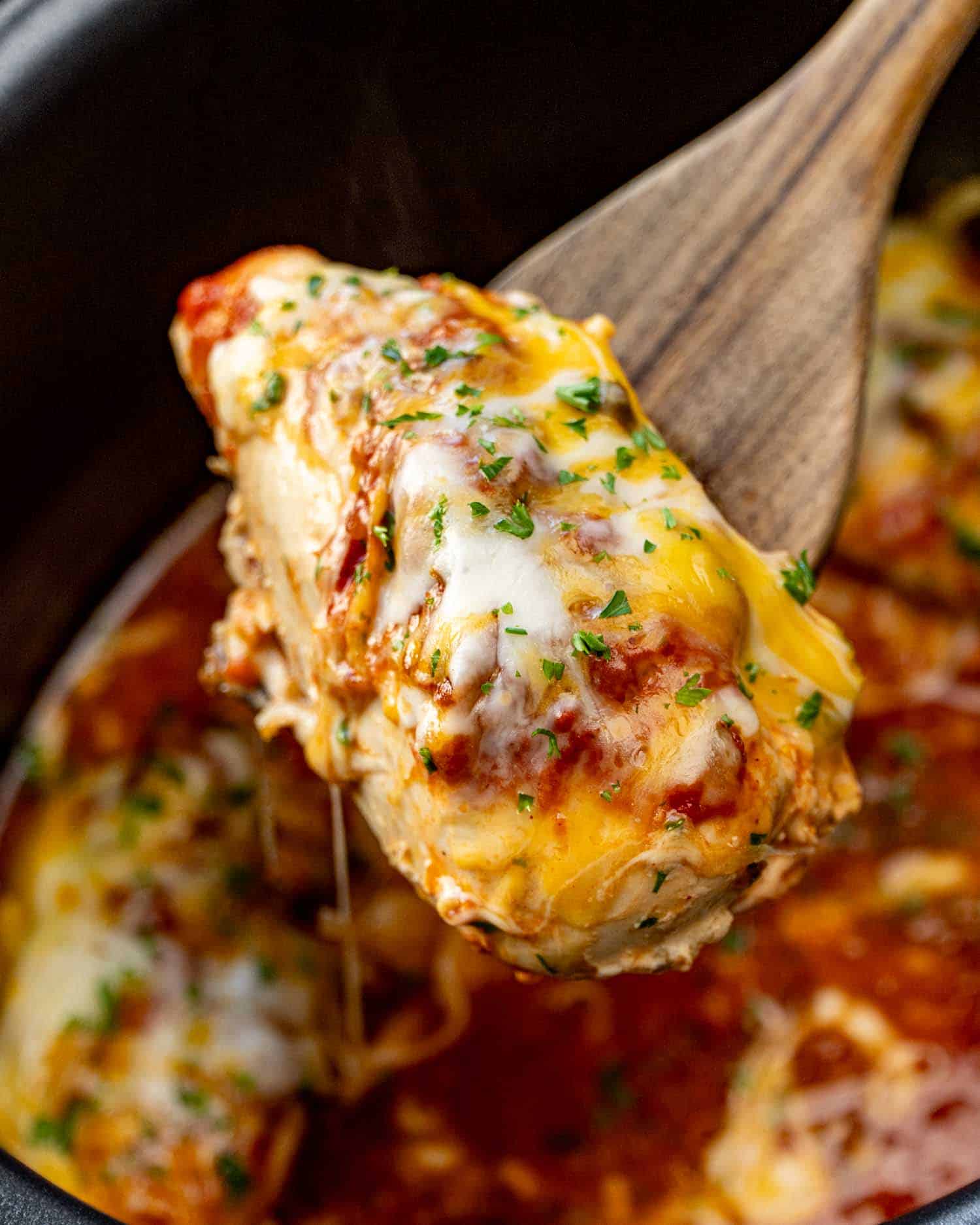 Crockpot salsa chicken breast lifted on a wooden spoon with gooey melted cheese, red salsa, and fresh parsley on top.
