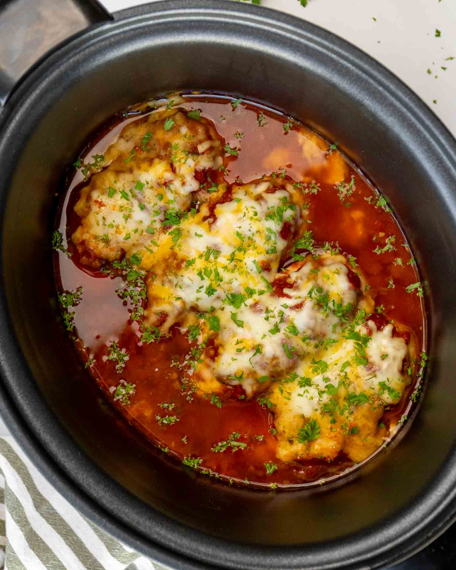 Overhead view of crockpot salsa chicken breasts simmering in red salsa, topped with melted cheese and freshly chopped parsley.