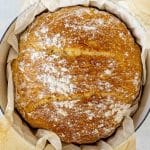 Top view of no knead bread in a Dutch oven lined with parchment, showing a rustic golden crust and cracks.