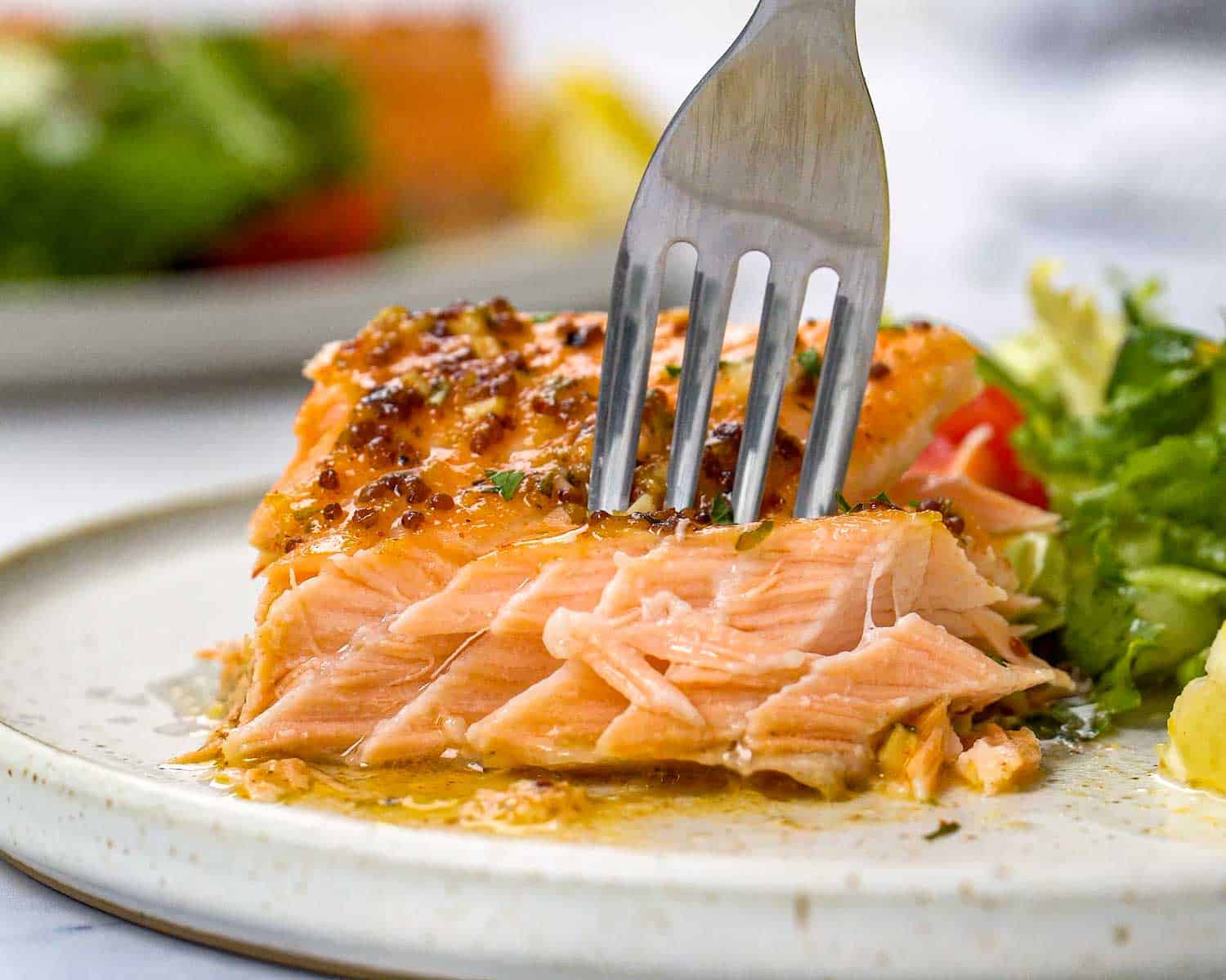 Fork cutting into baked honey mustard salmon showing tender flaky texture with buttery mustard sauce and fresh herbs.