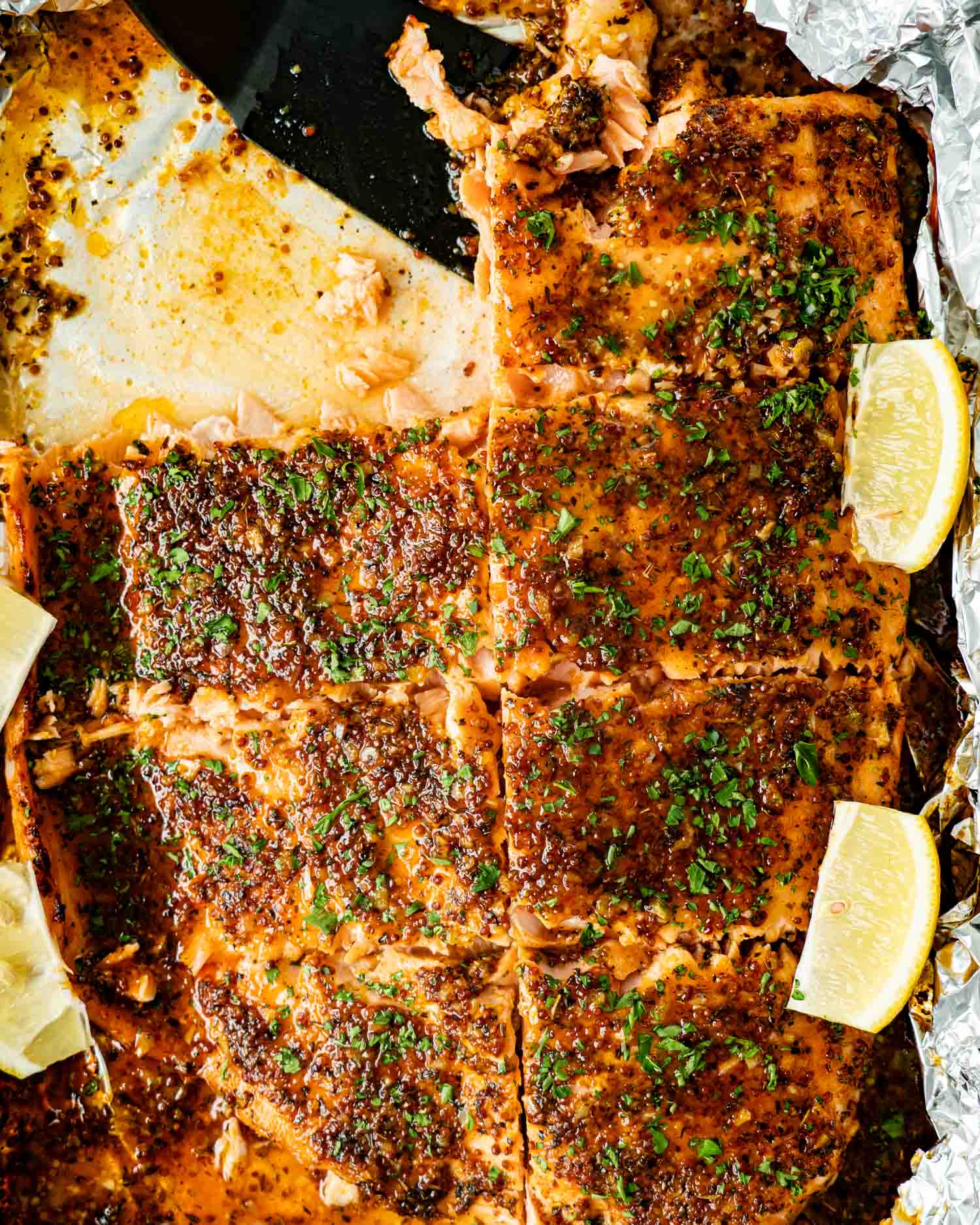 Baked honey mustard salmon cut into portions in foil pan with caramelized glaze, parsley, and lemon wedges for serving.