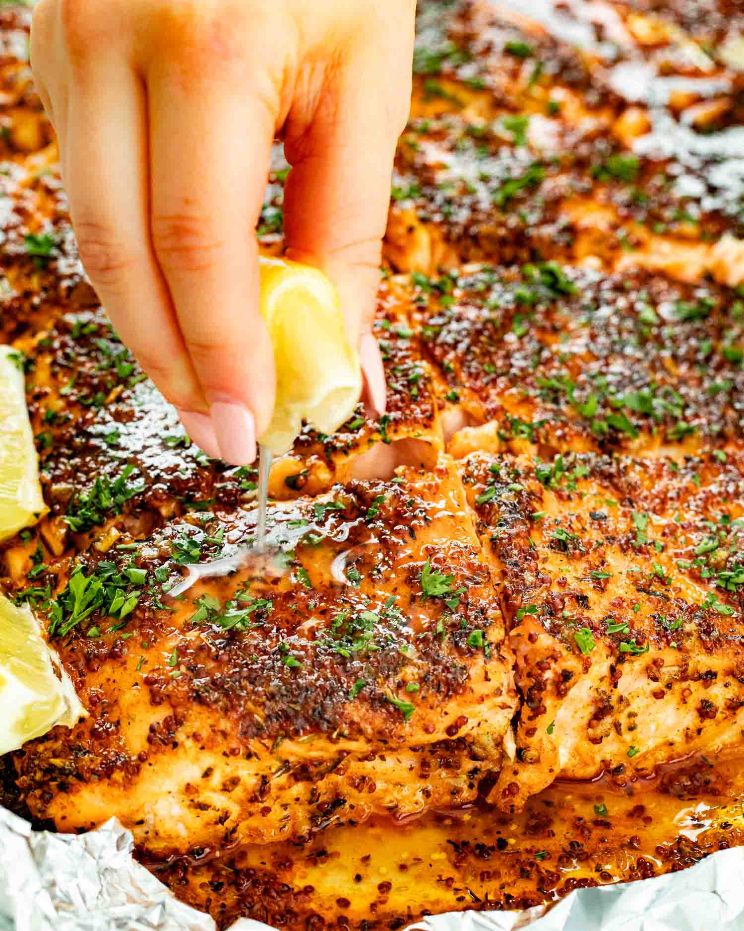Fresh lemon juice being squeezed over baked honey mustard salmon cooked in foil with herbs and caramelized mustard glaze.