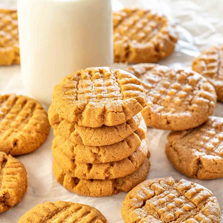 a stack of 5 peanut butter cookies with a glass of milk in the background surrounded by more cookies.