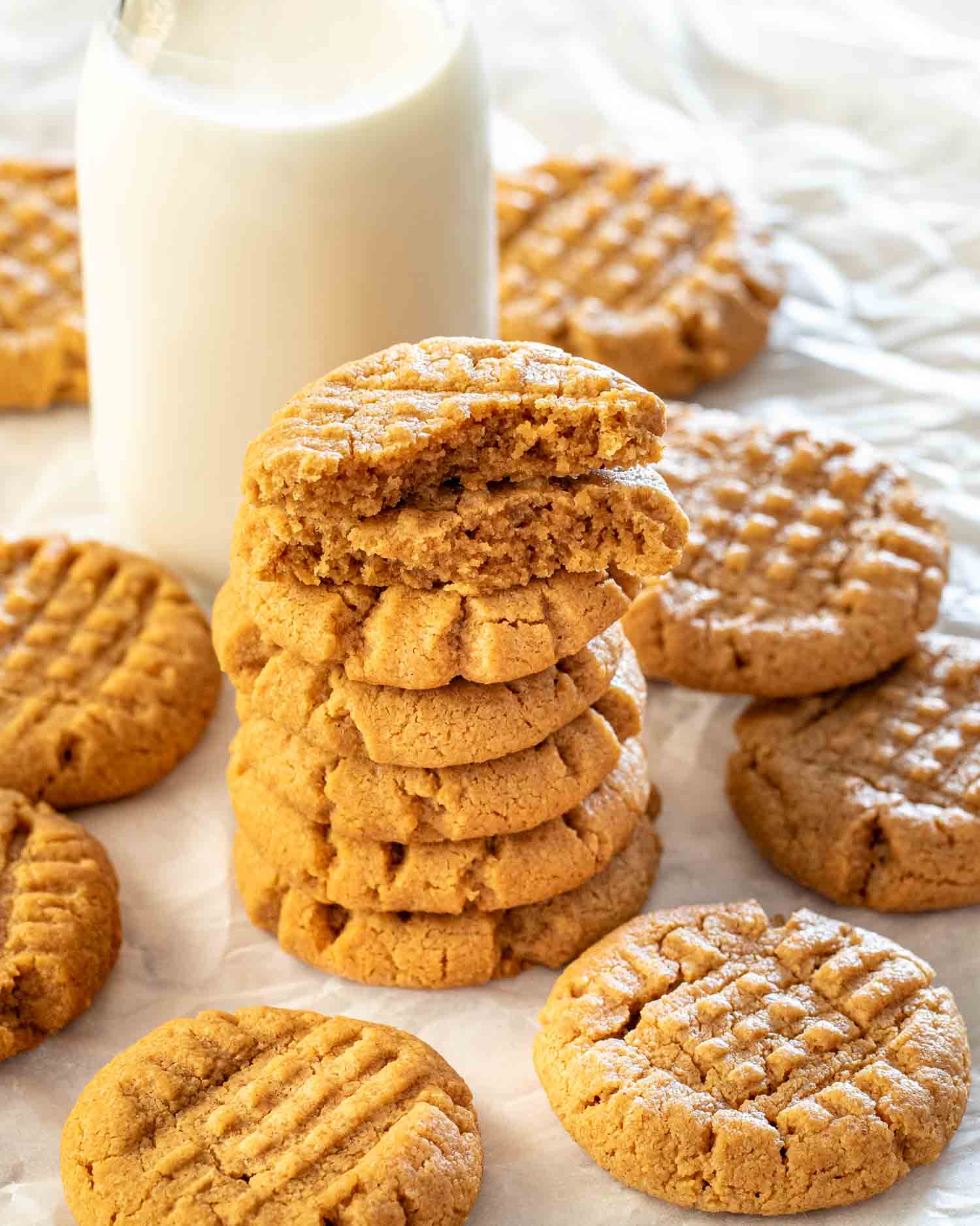 a stack of 5 peanut butter cookies with a glass of milk in the background surrounded by more cookies.