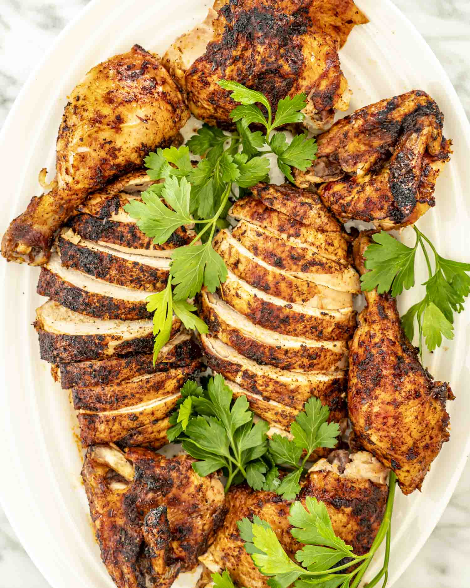 Sliced beer can chicken arranged on a platter with drumsticks, thighs, and fresh parsley for garnish.